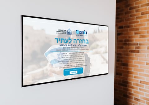 Flat screen monitor mounted on white wall next to brick, displaying Hebrew language website with blue design elements